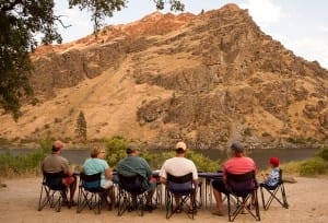 a family relaxes in the hells canyon river dining table looking out over the snake river for mountain goats or sheep on the steep rocky wall in front of them