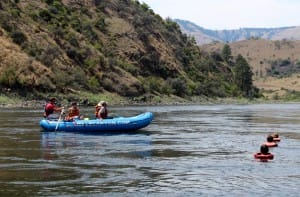 a boat floats downstream next to 3 people in the water cooling off from the hot summer heat on a salmon river rafting trip