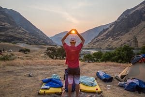 a man stands in a hells canyon camp with is arms above his head and his hands framing the setting sun