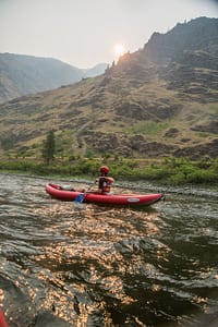 a kayaker in red floats down the snake river in the afternoon while an overnight rafting trip