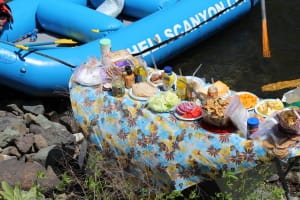 a full lunch spread of sandwiches, chips cookies and more in sitting on a table on a rocky beach on the snake river. a boat is waiting behind with hells canyon written on it