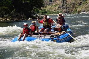 a guide smiles at the camera as the raft dips into a rapid with 4 kids ready to get splashed. Hells Canyon, Snake River rafting trip.