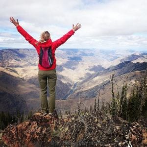 a backpacker with her hands in the air in celebration is on the edge of a cliff with the seven devils mountain range vista in front of her during a hell hike and raft hiking trip