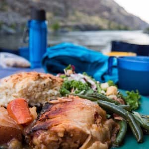 freshly baked bread, veggies and chicken are on a plate on a table with the snake river and hells canyon in the background on a whitewater rafting vacation